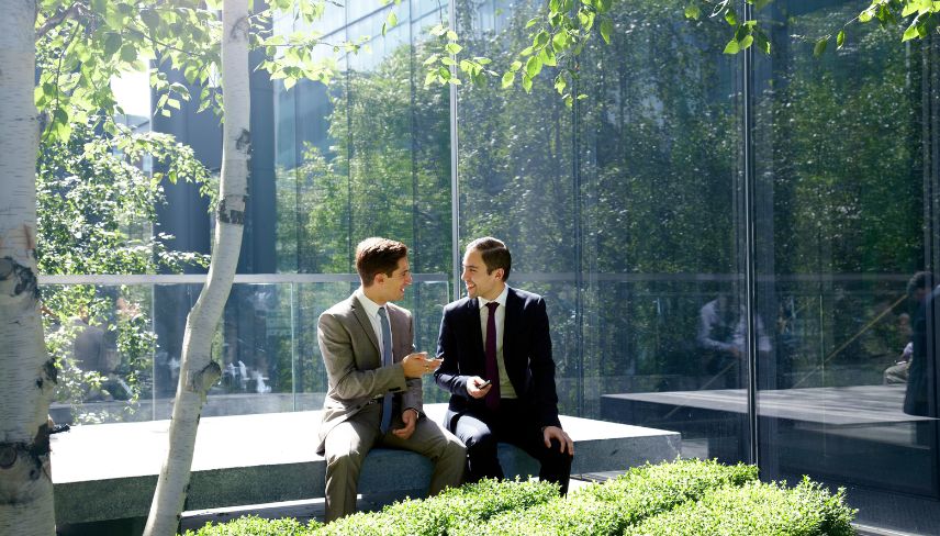 Two men in suits sit on a bench, facing a large glass wall, engaged in conversation.